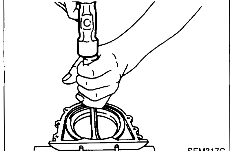 Hand using a screwdriver or pry tool to remove the old oil seal from the rear oil seal retainer that is sitting on a workbench.