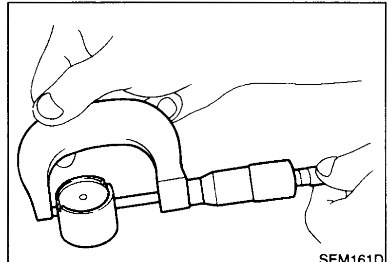 Hand using a micrometer to measure the outer diameter of a valve lifter held against the work surface.